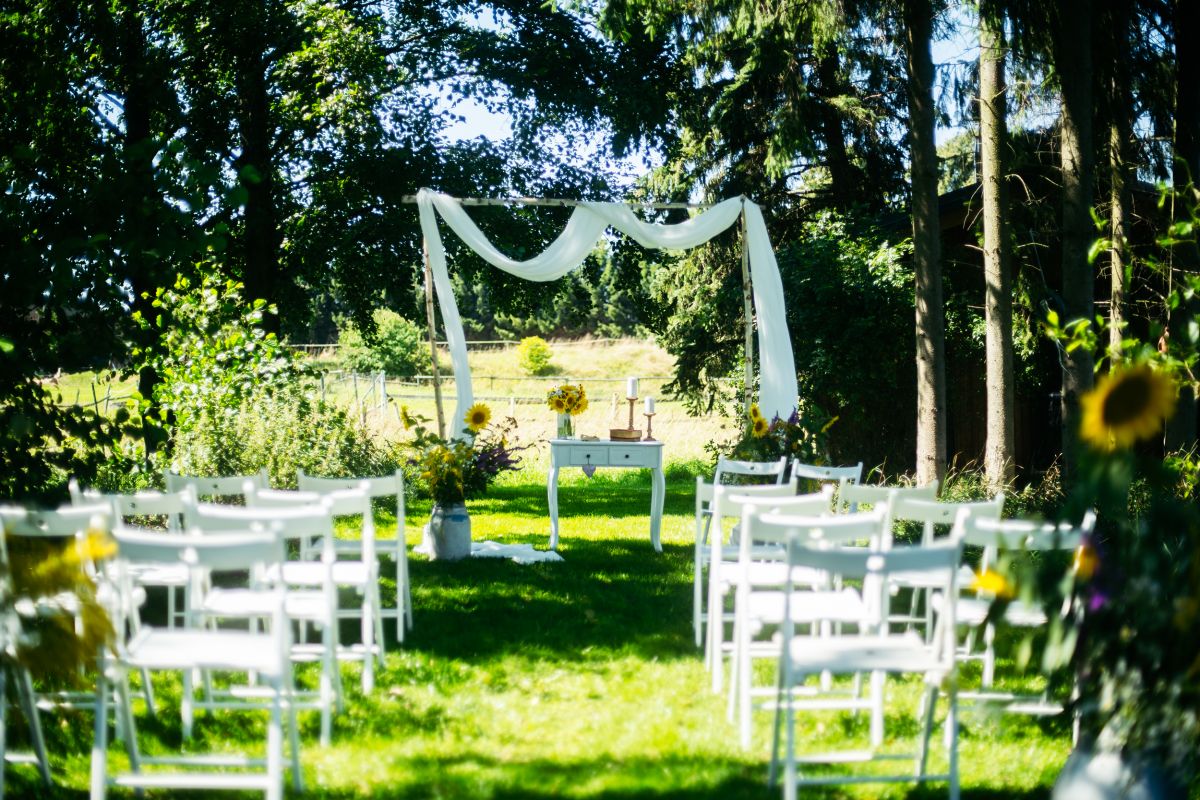 Traualter mit weißer Dekoration und Blumendeko auf einer grünen Wiese, davor weiße Holzstühle für die Hochzeitsgesellschaft