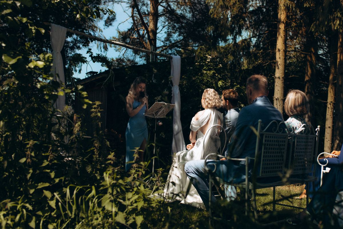 Hochzeitszeremonie im Grünen, Hochzeitspaar sitzt vor der Traurednerin inmitten von Bäumen