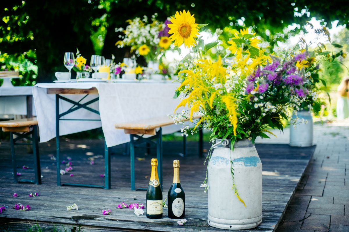 Ein großer, auf dem Boden stehender Blumenstrauß. daneben zwei Sektflaschen. Im Hintergrund eine Kaffeetafel im Garten.