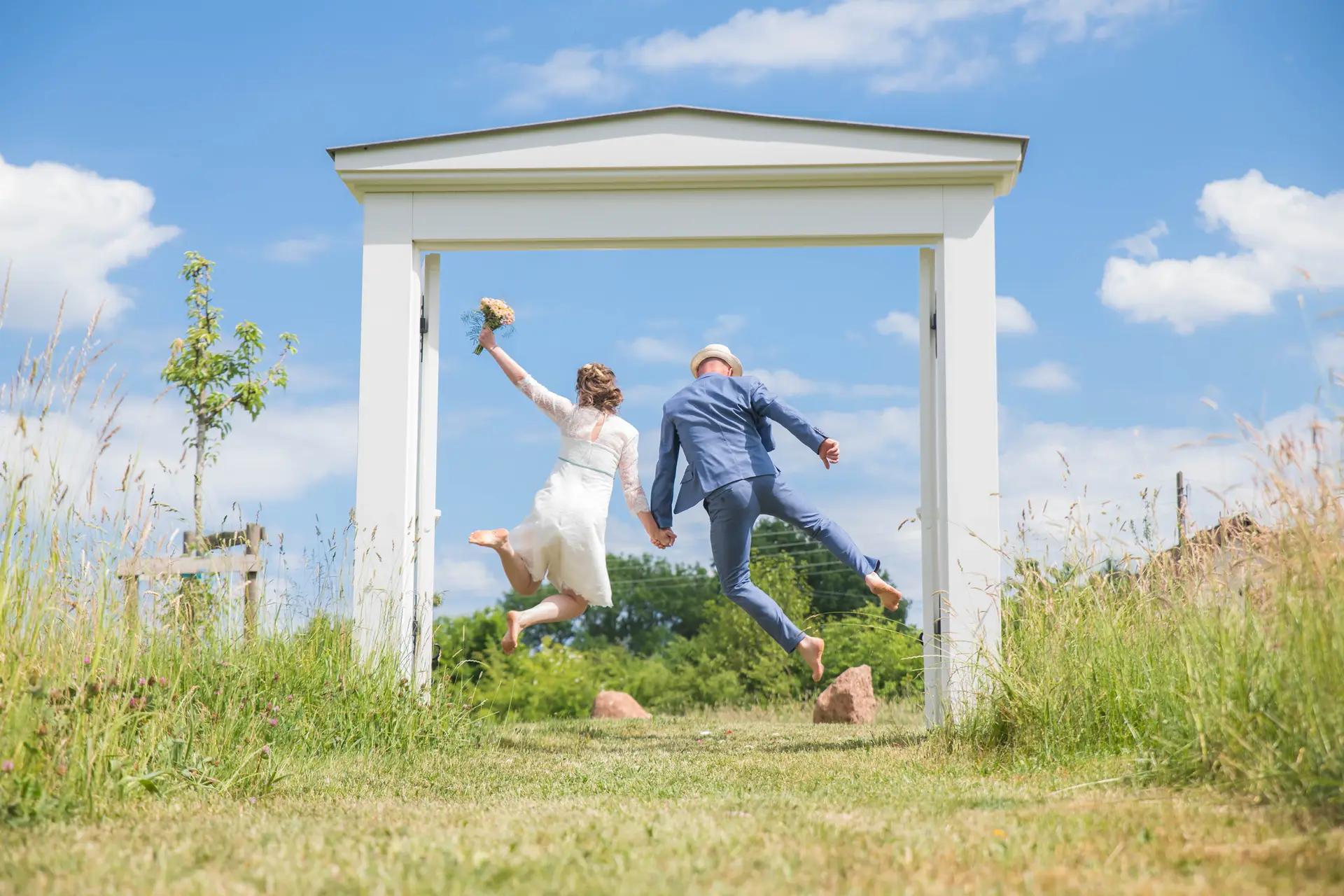 Von hinten fotografiertes Hochzeitspaar springt in einem weißen Holztor Hand in Hand in die Höhe.